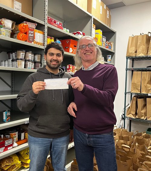 Two males standing in a room with shelves of food. One They're smiling and holding a cheque